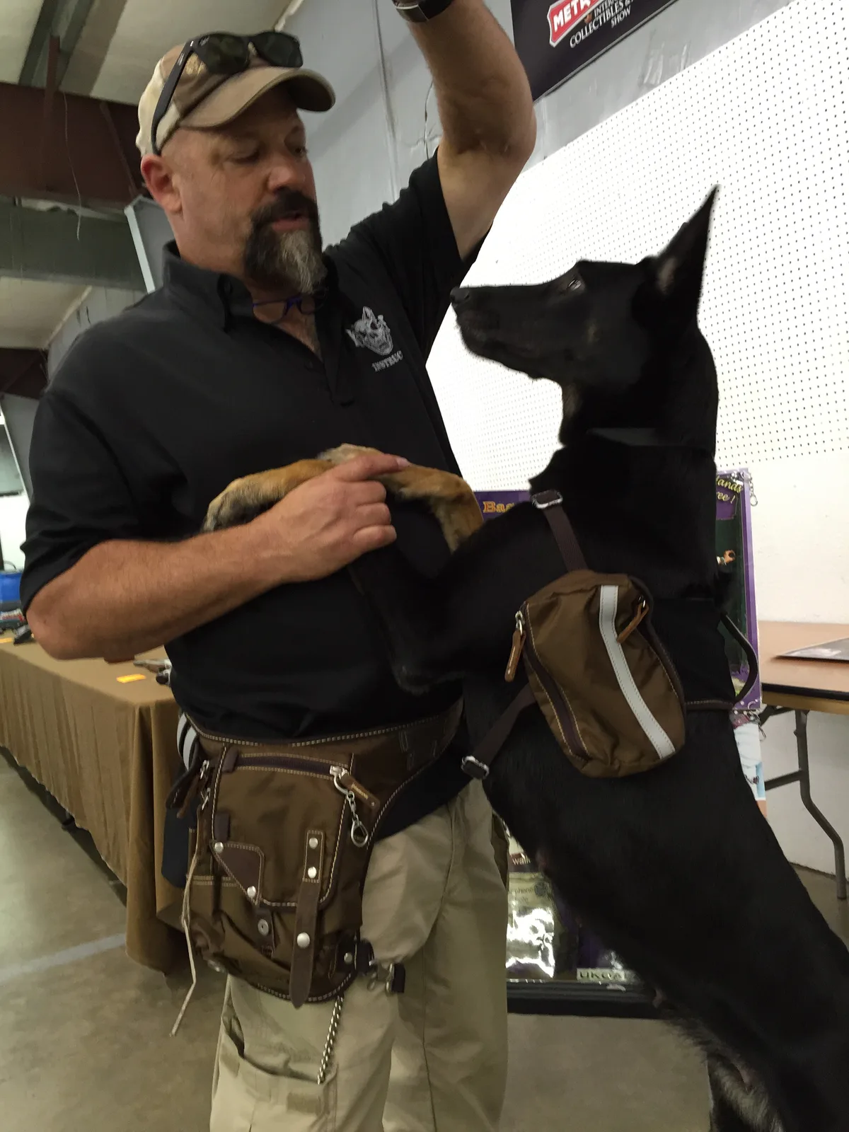 K9 handler at an expo wearing Ukoala bags on both hips while a black shepherd jumps up to greet him